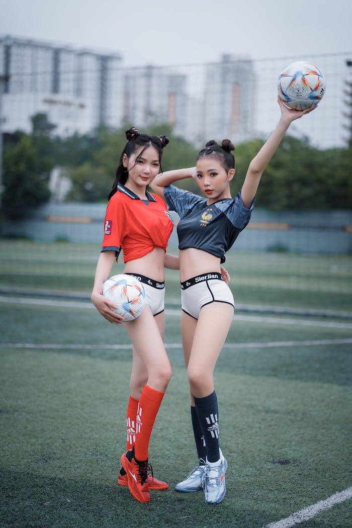 Two young women posing on a soccer field in colorful outfits, holding footballs.