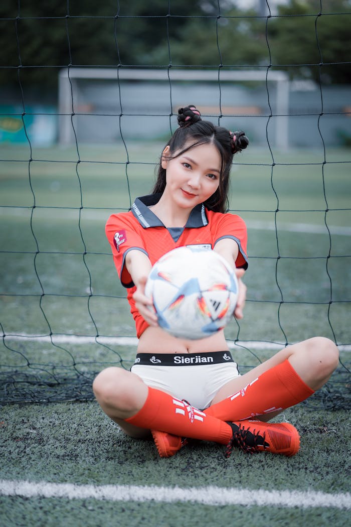 Asian woman in soccer uniform sitting on field holding a football.
